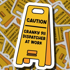 May include: A yellow A-frame caution sign sticker with a white border. The text reads CAUTION in black, followed by CRANKY 911 DISPATCHER AT WORK in black lettering. The sign features a handle cutout at the top and a rectangular graphic detail.