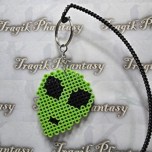 May include: A green and black alien face keychain made of small, round beads. The alien has large black eyes and a smiling mouth. The keychain is attached to a silver keyring and a black cord.