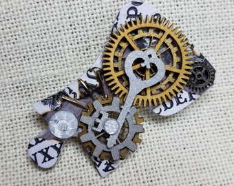 May include: A silver and gold steampunk-style puzzle piece brooch with a key and gears. The piece is made of paper and has a distressed look.