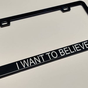 A photo showing a black license plate frame with white text saying I want to believe in thin helvetica font