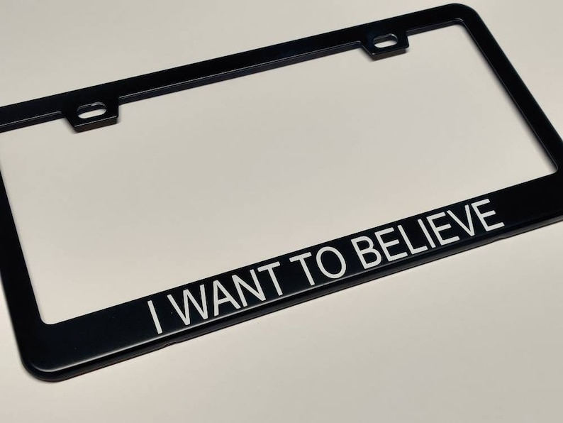 A photo showing a black license plate frame with white text saying I want to believe in thin helvetica font