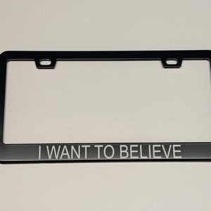 A photo showing a black license plate frame with white text saying I want to believe in thin helvetica font