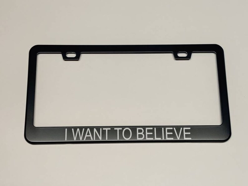 A photo showing a black license plate frame with white text saying I want to believe in thin helvetica font