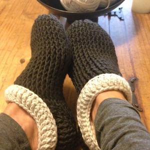 Puede incluir: Un par de zapatillas acogedoras de punto gris oscuro con ribete de ganchillo blanco. Las zapatillas están puestas en los pies de una persona.
