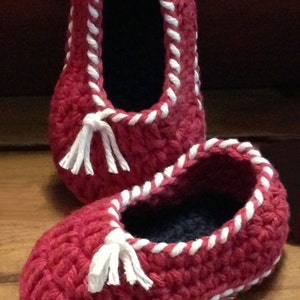 May include: A pair of red crocheted slippers with white trim and tassels. The slippers are made with a chunky yarn and have a simple, classic design.