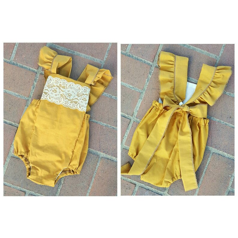 May include: A mustard yellow baby onesie with a white lace trim and ruffled straps. The onesie has a bow tie in the back.