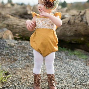 May include: A young girl wearing a mustard yellow romper with white lace trim and white tights. She is wearing brown boots and has a flower headband on her head.