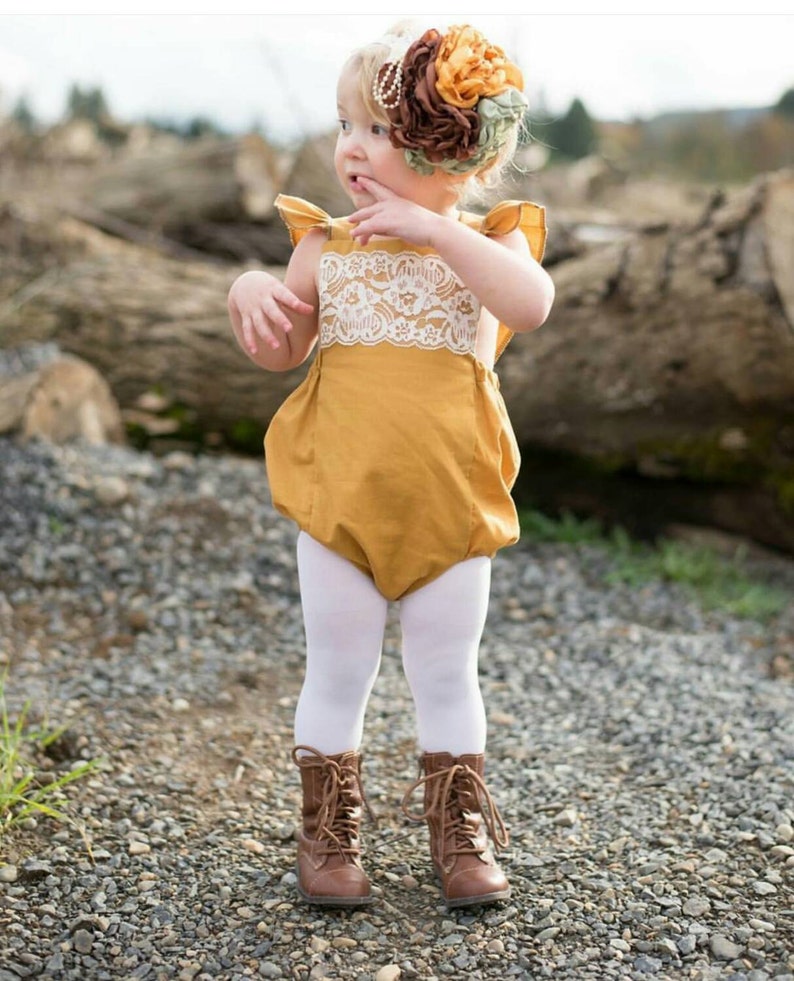 May include: A young girl wearing a mustard yellow romper with white lace trim and white tights. She is wearing brown boots and has a flower headband on her head.