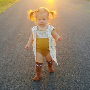 May include: A young child wearing a mustard yellow bodysuit with a white lace overlay and brown knee-high socks with a fox design. The child has pigtails and is walking on a paved road.