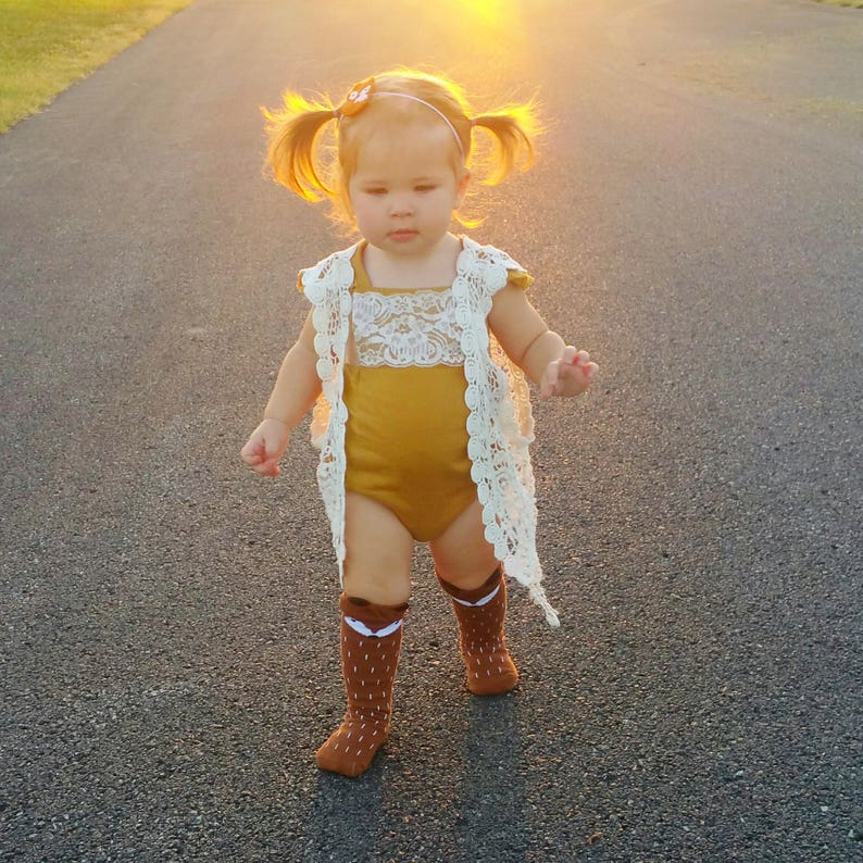 May include: A young child wearing a mustard yellow bodysuit with a white lace overlay and brown knee-high socks with a fox design. The child has pigtails and is walking on a paved road.
