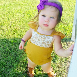 May include: A little girl wearing a mustard yellow romper with a white lace trim and a purple bow headband. She is standing on green grass and wearing brown sandals.
