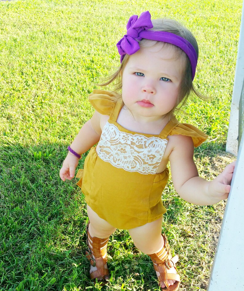 May include: A little girl wearing a mustard yellow romper with a white lace trim and a purple bow headband. She is standing on green grass and wearing brown sandals.