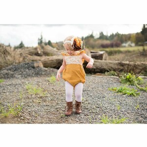 May include: A little girl wearing a mustard yellow romper with white lace trim and white tights. She is wearing brown boots and a flower headband.