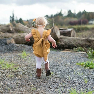May include: A little girl wearing a mustard yellow romper with a bow in the back, white tights, and brown boots. She is walking away from the camera on a gravel path.