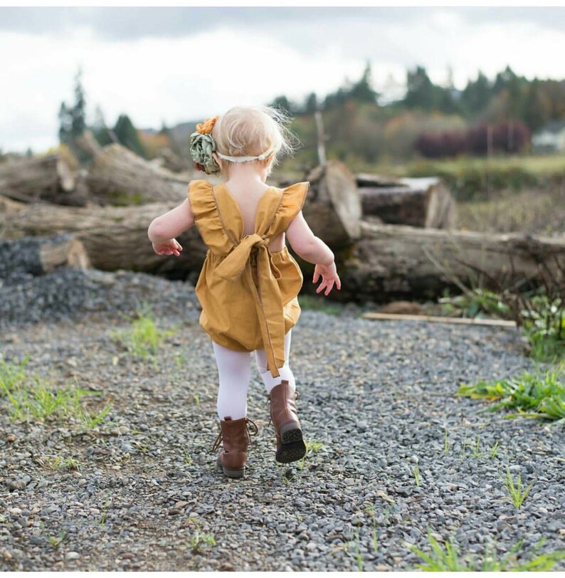 May include: A little girl wearing a mustard yellow romper with a bow in the back, white tights, and brown boots. She is walking away from the camera on a gravel path.