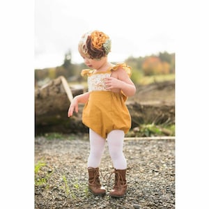 May include: A little girl wearing a mustard yellow romper with white lace trim and white tights. She is wearing brown lace-up boots and a flower headband.