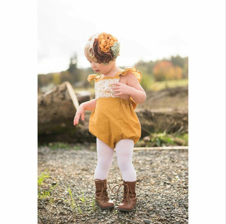 May include: A little girl wearing a mustard yellow romper with white lace trim and white tights. She is wearing brown lace-up boots and a flower headband.