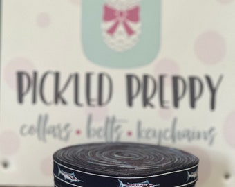 May include: A roll of navy blue ribbon with a white marlin fish design. The ribbon is labeled "PICKLED PREPPY collars - belts - keychains".