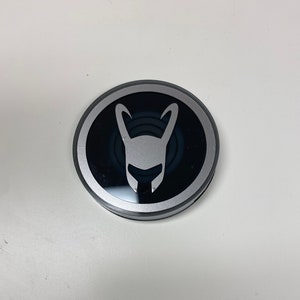 May include: A round, silver-rimmed button featuring a black center with a white, stylized horned helmet design. The button has a metallic silver border and a glossy black center, set against a white background.