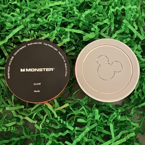 May include: A black and red circular cup holder light with the text "MONSTER" and "On/Off Mode" printed on it. The light is next to a silver cup holder with a Mickey Mouse silhouette.