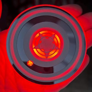 May include: A round, gray and black device with a red glowing star design in the center. The device has concentric circles and a red illuminated edge. A small, red light is also visible.