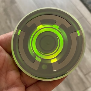 Op de afbeelding: Een ronde, zilverkleurige sticker met een concentrisch cirkelontwerp in groen en geel. De sticker heeft een glanzende afwerking.