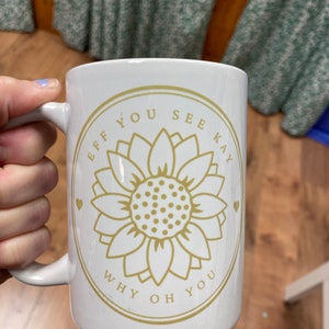 以下が含まれることがあります： 白いセラミック製のマグカップで、金色の日向葵のデザインと「Eff You See Kay Why Oh You」という文字が書かれています。