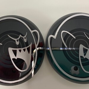 May include: Two black and white round stickers with a ghost design. The ghost is outlined in white and has a swirling pattern inside. The ghost has a large, toothy grin and two eyes.