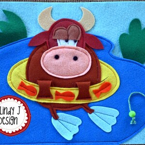 Puede incluir: Un toro de fieltro marr&oacute;n con un salvavidas amarillo y aletas azules est&aacute; nadando en una piscina de fieltro azul. El toro tiene una nariz rosa y ojos marrones. El salvavidas tiene peces naranjas en &eacute;l. La piscina tiene plantas de fieltro verde en los lados. El texto "Lindy J Design" est&aacute; en un c&iacute;rculo de fieltro rojo.