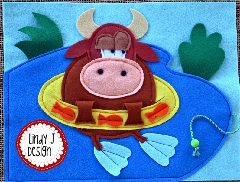 Puede incluir: Un toro de fieltro marr&oacute;n con un salvavidas amarillo y aletas azules est&aacute; nadando en una piscina de fieltro azul. El toro tiene una nariz rosa y ojos marrones. El salvavidas tiene peces naranjas en &eacute;l. La piscina tiene plantas de fieltro verde en los lados. El texto "Lindy J Design" est&aacute; en un c&iacute;rculo de fieltro rojo.