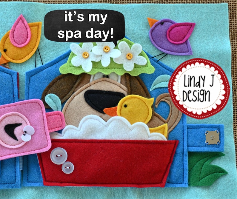 Puede incluir: Una p&aacute;gina de libro de fieltro con un perro tomando un ba&ntilde;o. El perro lleva una corona de flores y est&aacute; en una ba&ntilde;era roja con espuma blanca. El texto "it's my spa day!" est&aacute; escrito en una pizarra negra. El texto "Lindy J Design" est&aacute; escrito en un c&iacute;rculo rojo.