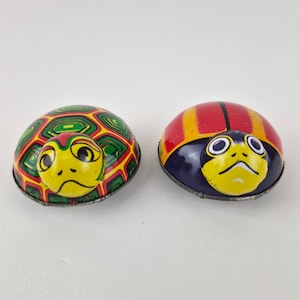 May include: Two small metal wind-up toys shaped like creatures. The left toy features a green and yellow geometric shell pattern with a yellow head. The right toy has a red and yellow striped shell with a dark blue base and a yellow head.