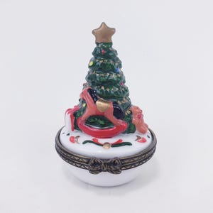 May include: A white, oval-shaped trinket box with a Christmas tree design. The tree is green with gold star, and the box features a rocking horse, gifts, and a bow. The box has a bronze-colored trim.