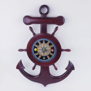 May include: A nautical-themed wall clock in the shape of an anchor. The clock face features a compass design with a rope border and billiard ball accents. The clock is made of dark brown wood and has a ship's wheel design.