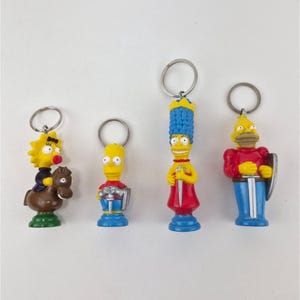 May include: Four keychains featuring The Simpsons characters in medieval attire. Maggie rides a brown horse, Bart holds a sword, Marge wears a crown and holds a sword, and Homer wears red armor with a shield. Each figure has a silver metal ring attached.
