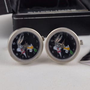 May include: A pair of silver-toned cufflinks featuring a black watch face with white numerals and Bugs Bunny holding a carrot. The cartoon character is in color, with a red bow tie and green accents.