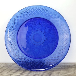 May include: A vibrant blue glass plate with a diamond pattern around the rim and a starburst design in the center. The plate is round and appears to be vintage, with a deep, rich color. The plate is likely used for serving or display.
