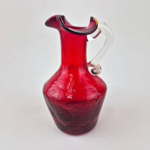 May include: A small, vibrant red glass pitcher featuring a distinct crackle texture throughout the body. The vessel has a wide, ruffled rim and a clear, curved glass handle attached to the side, creating a contrast with the deep red glass.