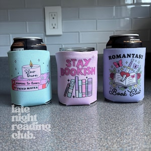 May include: Three beverage insulators with book-themed designs. One is light blue with a candle and the text "Slow Burn". Another is pink with "Stay Bookish". The third is lavender with "Romantasy Book Club".