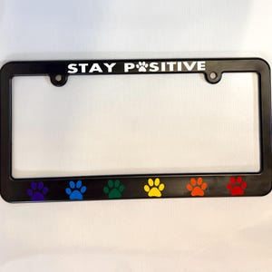 May include: Black license plate frame with the text "STAY POSITIVE" in white lettering with a paw print symbol. The bottom of the frame has six rainbow paw prints.