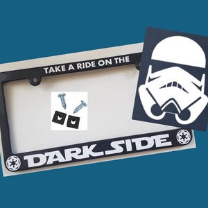 May include: Black license plate frame with white text that reads "Take a ride on the dark side" and a Star Wars symbol. The frame includes two black plastic screw covers and two silver screws. A separate sticker of a white Stormtrooper helmet is included.