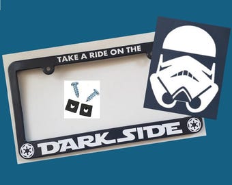 May include: A black license plate frame featuring the white text TAKE A RIDE ON THE DARK SIDE and two Imperial crest icons. Included are two silver metal screws and two small black square plastic fasteners for mounting the frame to a vehicle.