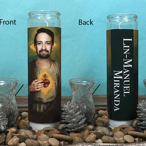 May include: A white candle with a picture of Lin-Manuel Miranda on the front and his name on the back. The candle is sitting on a bed of small rocks.
