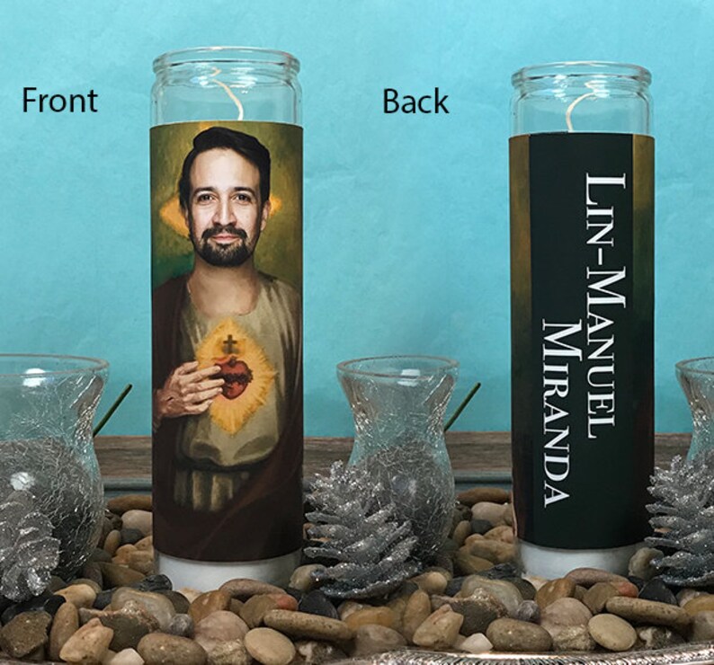 May include: A white candle with a picture of Lin-Manuel Miranda on the front and his name on the back. The candle is sitting on a bed of small rocks.