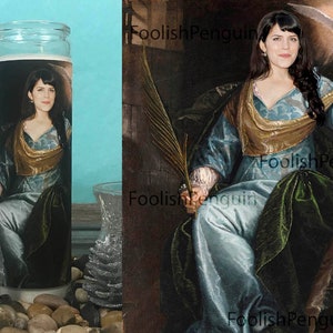 May include: A white candle with a printed image of a woman in a blue and gold dress holding a white bird. The woman has long dark hair and is wearing a gold crown.