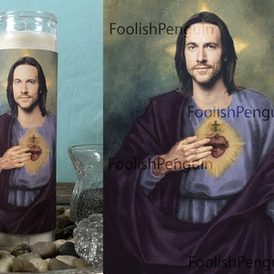 May include: A white candle with a printed image of a man with long brown hair, wearing a purple robe and holding a heart with a crown of thorns and a cross. The image is surrounded by a gold halo.