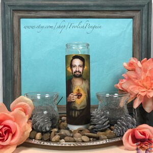May include: A white candle with a printed image of a man with a beard and a heart on his chest. The candle is on a silver tray with small rocks and pine cones around it. The text "www.etsy.com/shop/FoolishPenguin" is visible in the background.