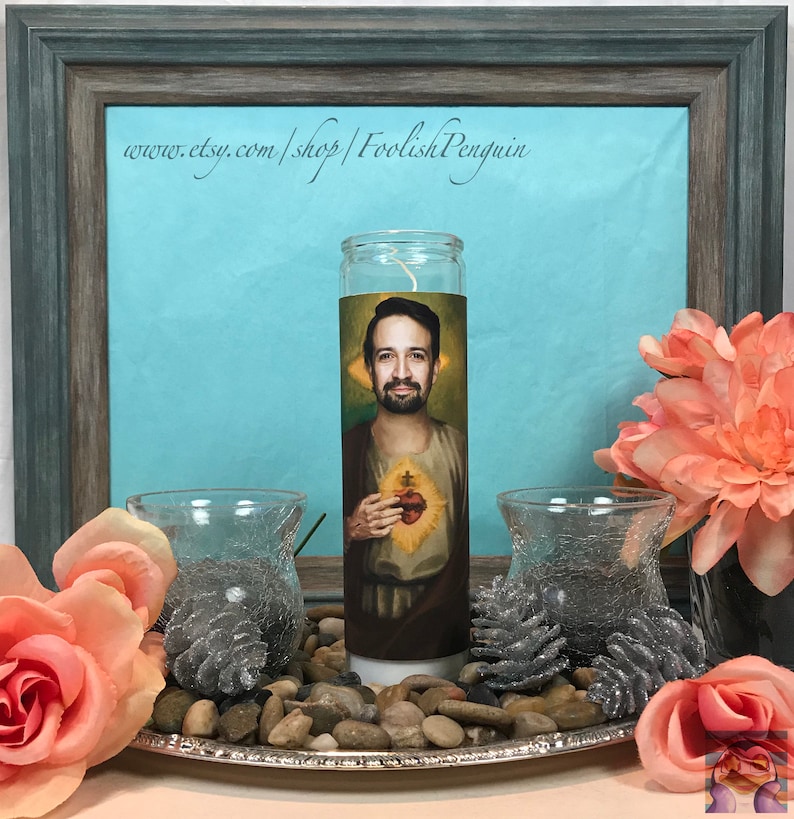 May include: A white candle with a printed image of a man with a beard and a heart on his chest. The candle is on a silver tray with small rocks and pine cones around it. The text "www.etsy.com/shop/FoolishPenguin" is visible in the background.