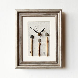 May include: A framed clock with a grey background and a white mount. The clock face is made of three small stones and twigs. The clock hands are black.