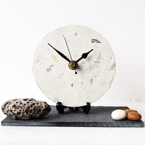May include: A circular clock with a textured, off-white face, black hands, and a brass centre. It rests on a black stand on a dark grey rectangular base, with a rock and small stones. The clock face has a natural, organic appearance, suggesting a handmade design.